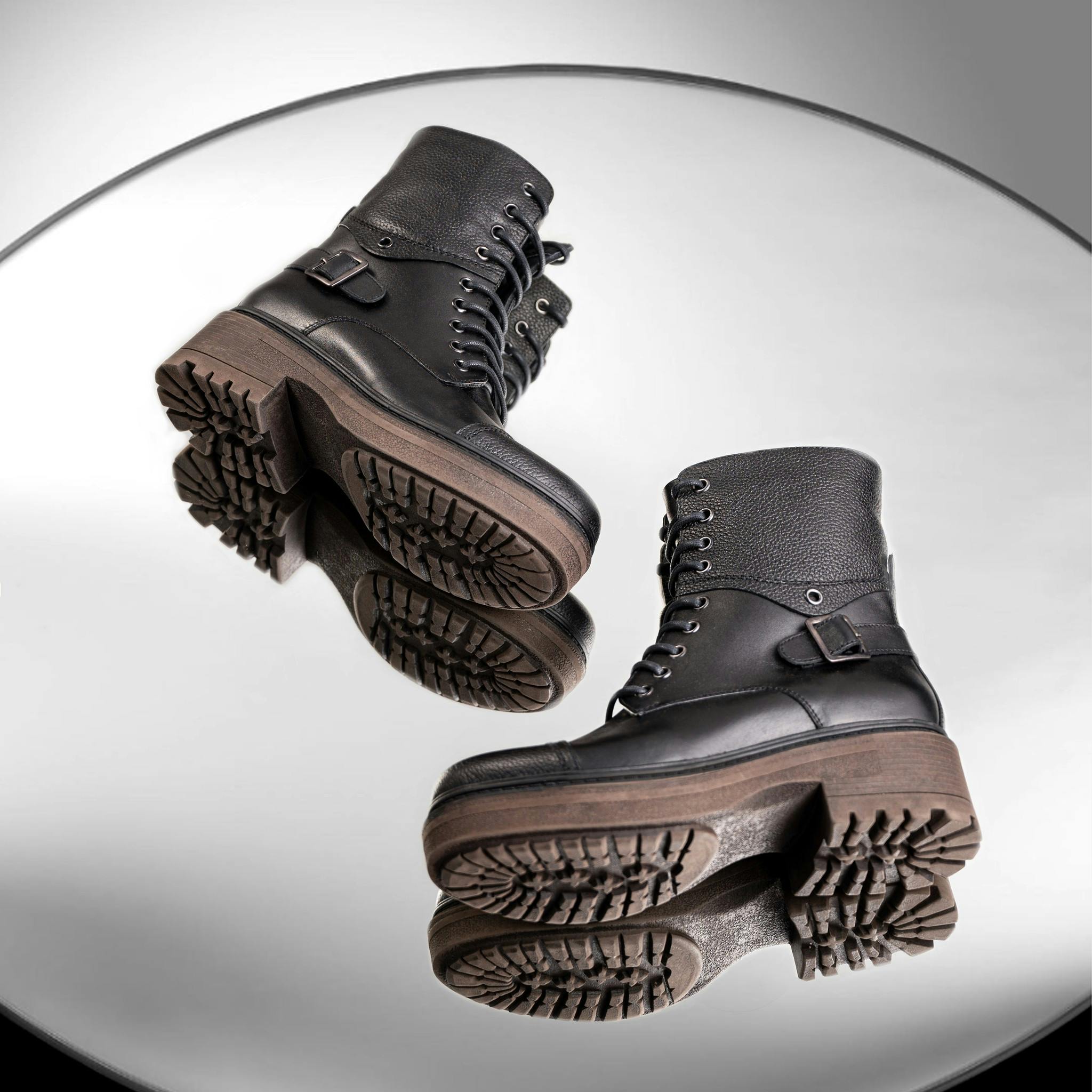 Black leather combat boots with treaded soles and reflective effect on a mirror surface.
