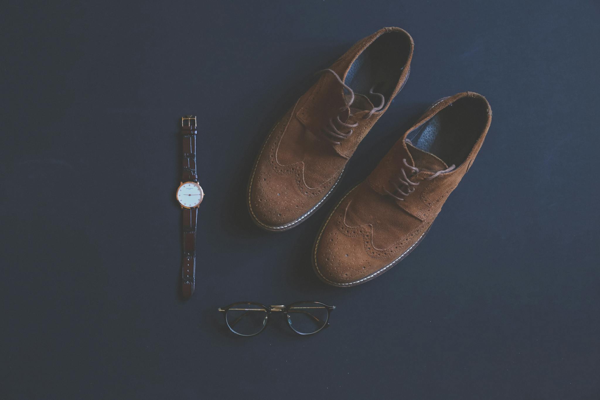 Flat lay of brown leather shoes, eyeglasses, and wristwatch on a dark background.