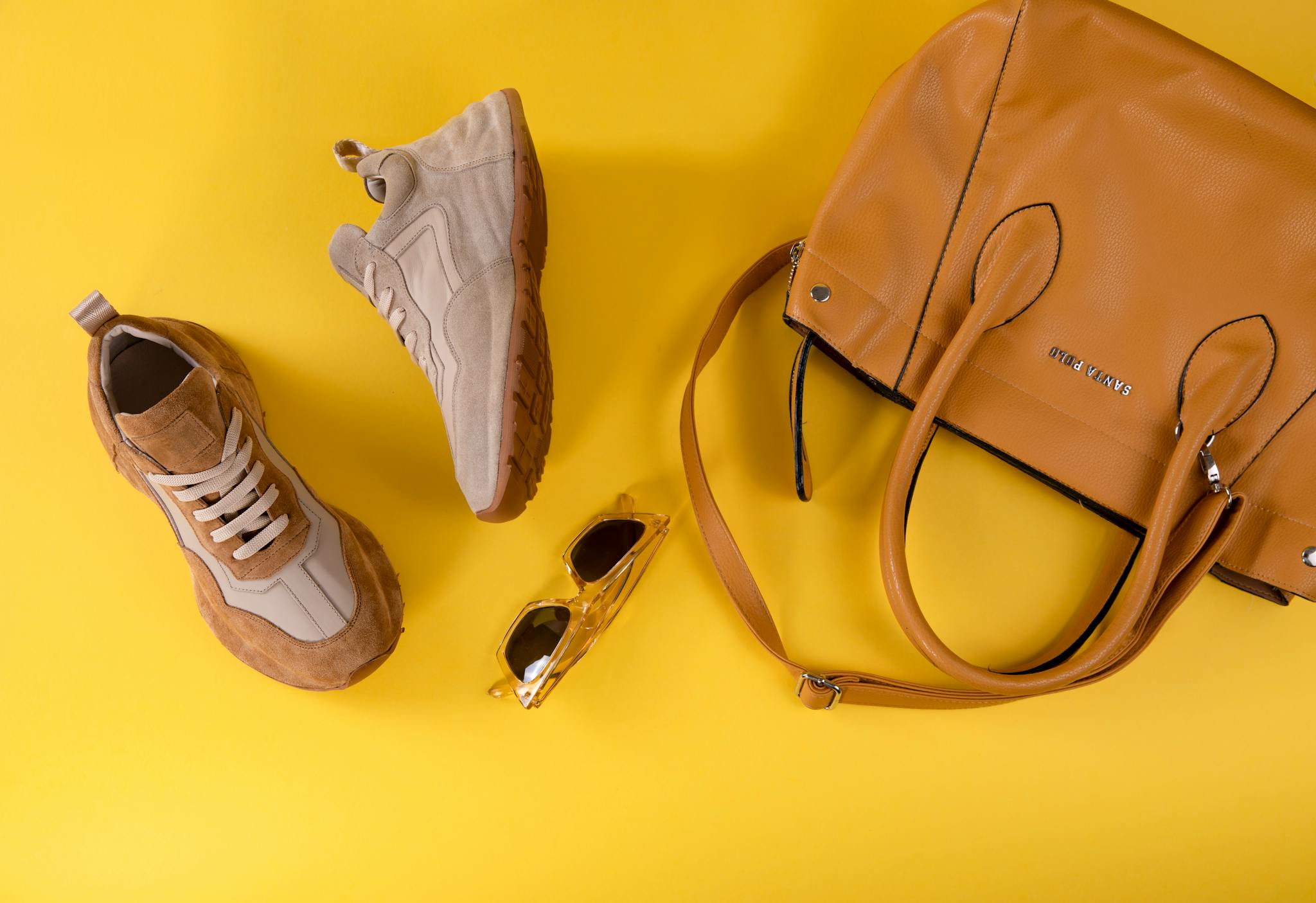 Stylish brown handbag, fashionable eyewear, and sport shoes on vibrant yellow background.